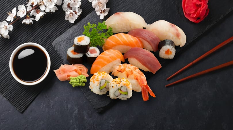 Assorted sushi and sashimi platter with soy sauce, ginger, wasabi, and chopsticks on a dark mat.