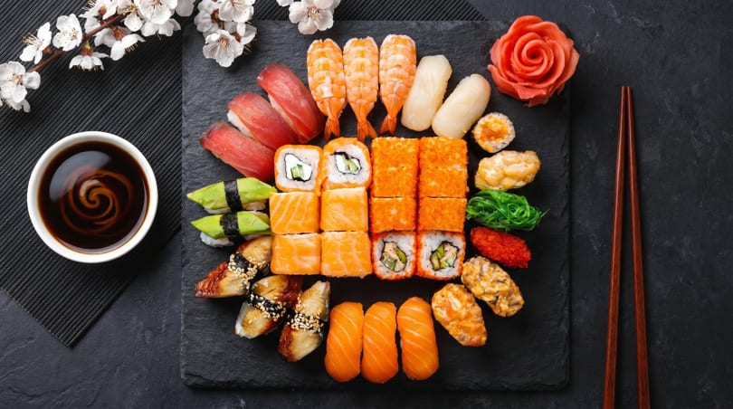 Assorted sushi and sashimi platter with soy sauce, chopsticks, and cherry blossoms on a dark slate.
