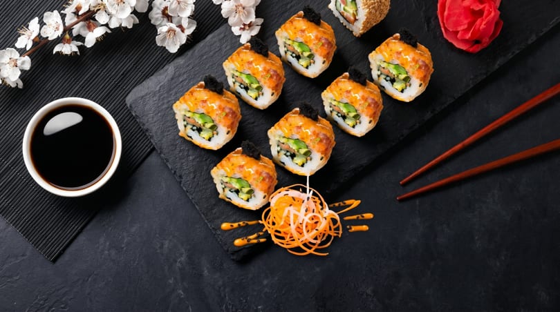 Top-down view of sushi rolls with orange sauce and black caviar, soy sauce, and chopsticks.